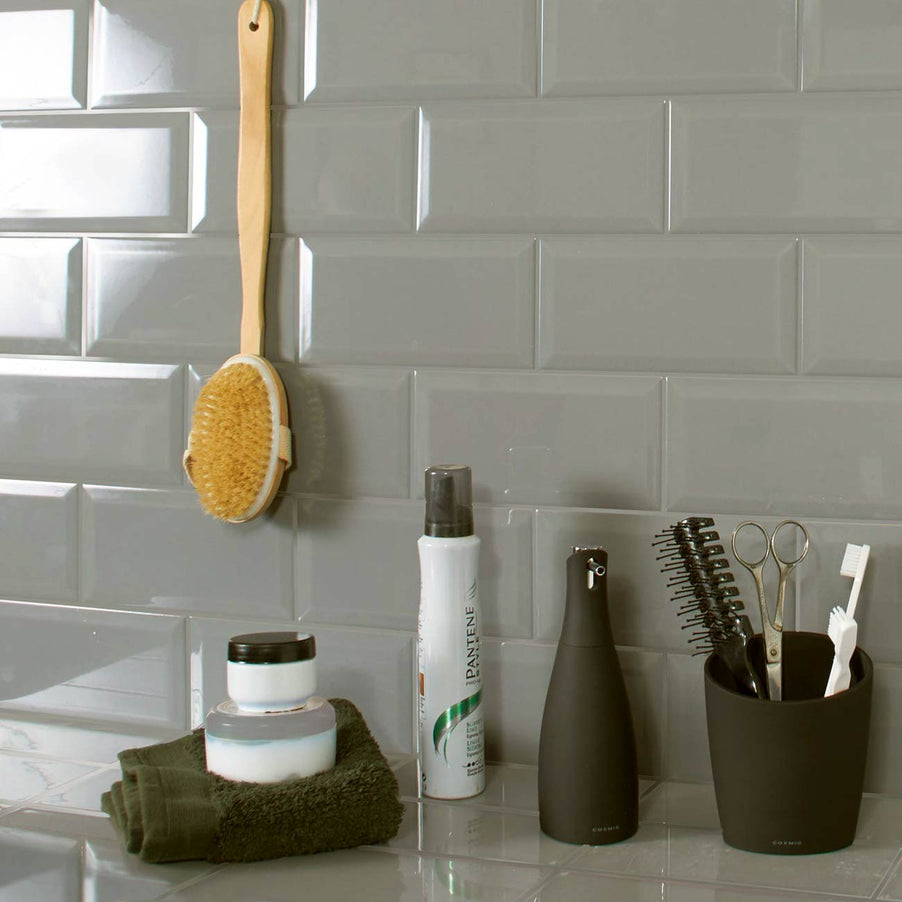 Metro Grey Plata Gloss Bevelled Brick tiles 10x20cm. Grey brick tiles inn a bathroom setting with a shelf featuring hair products, toothbrush and toothpaste. Also with a wooden body brush on the wall.