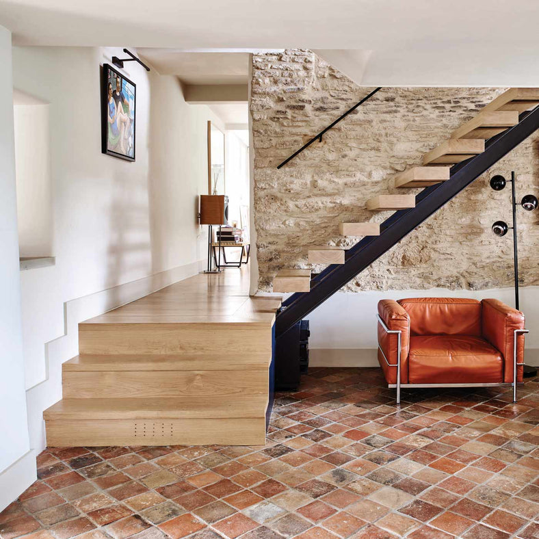 Antique Burgundy Terracotta Tiles 15x15cm by Ca' Pietra in a hallway setting. Satirs leading up and round with red armchair and lamp to the right. Cream painted walls and bare stone feature wall up the stairs.