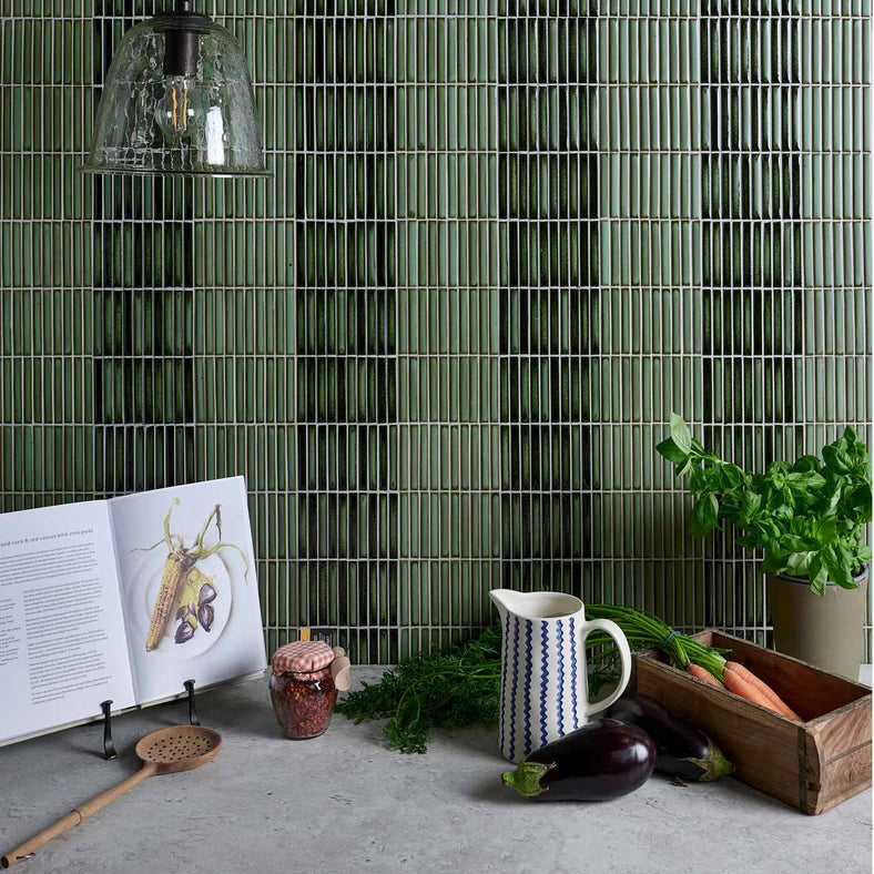 Bamboo Lustre Forest Satin Mosaic 28.2x29.4cm. Green mosaic tiles as kitchen splashback tiles above white stone effect worktop laden with cookkbook, carrots, potted basil, aubergine, wooden slotted spoon, and jug. Glass vintage pendant light hanging down.