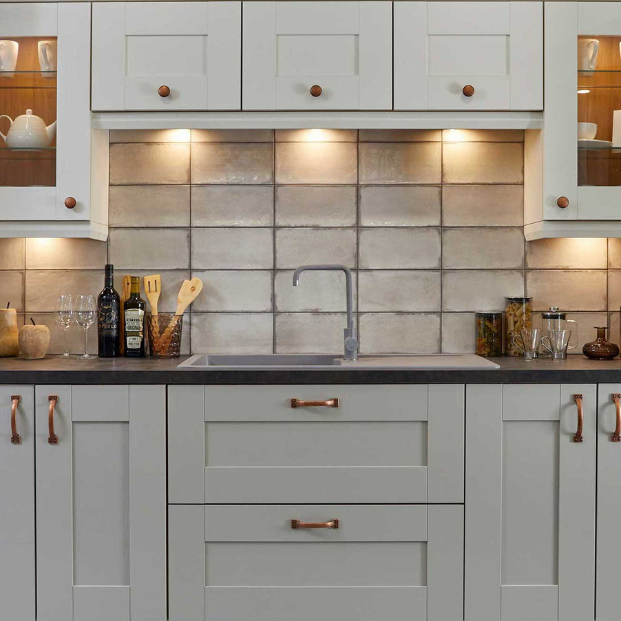 Essence Blanco tile 15x30cm white kitchen tiles and gloss tiles in a kitchen setting with light grey kitchen cabinets, dark worktop. Wall units to the top with under cabinet lighting and glasses and wooden cutlery placed on worktop next to the sink.