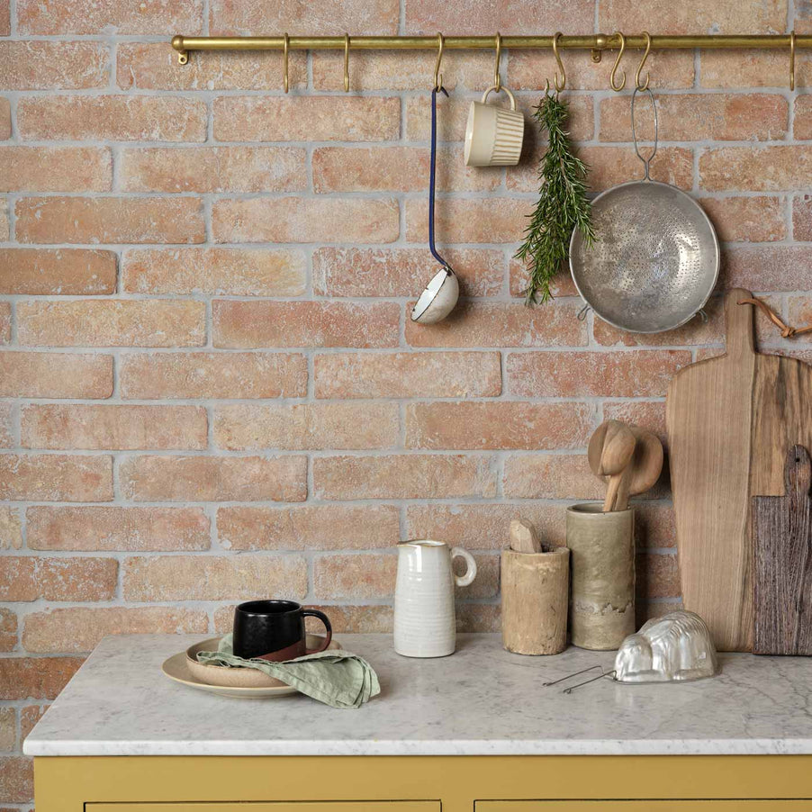 Marlborough Parquet Terracotta Tiles 7.5x30.5cm. Terracotta kitchen wall and terracotta brick tiles. In a kitchen setting with yellow painted cabinets and white stone worktop. Brass shelf with utensils hanging down. Wooden chopping boards on the worksurface.