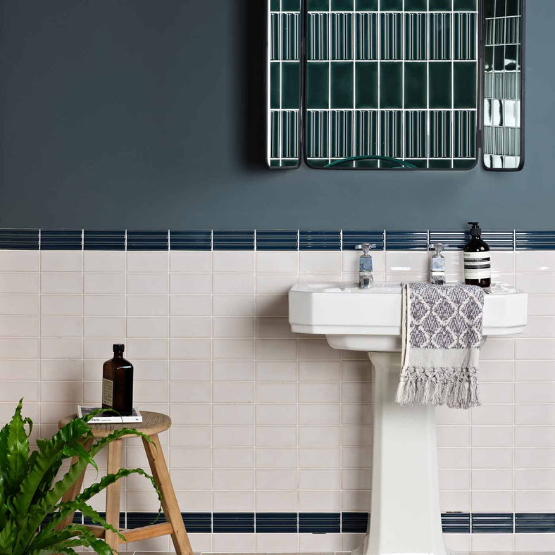 Tunstall Deep White Brick tiles 6.2x12.5cm in a bathroom setting with blue decor tiles in the middle, white pedestal sink, dark blue paint above the tiles. Features a wall mirror and a wooden stool with potted plant on top.