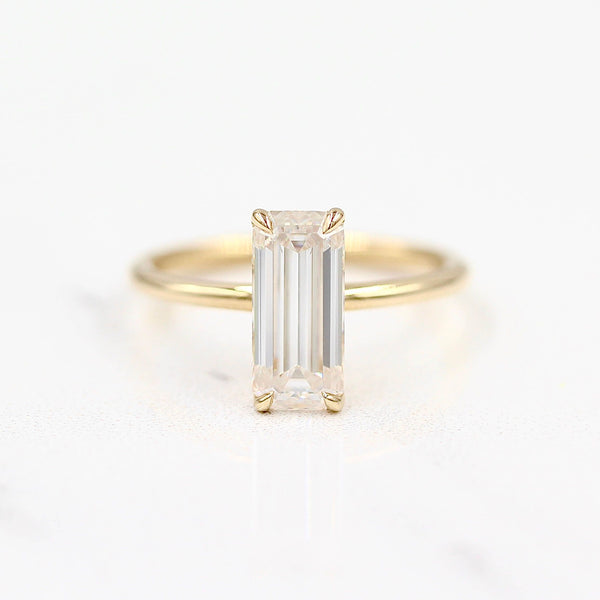 Solitaire elongated emerald diamond on a gold band on a reflective white surface