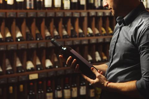 person holding standard size wine bottle in front of shelf of wine bottles