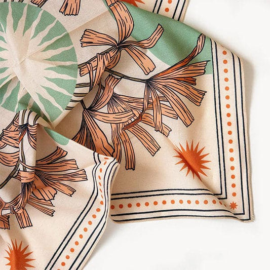 This soft peach cotton bandana is filled with a floral design of orange, long petaled flowers and is bordered by dark orange circles and black line work. In the center sits a silhouette of a beaming sun within a pastel green circle. 