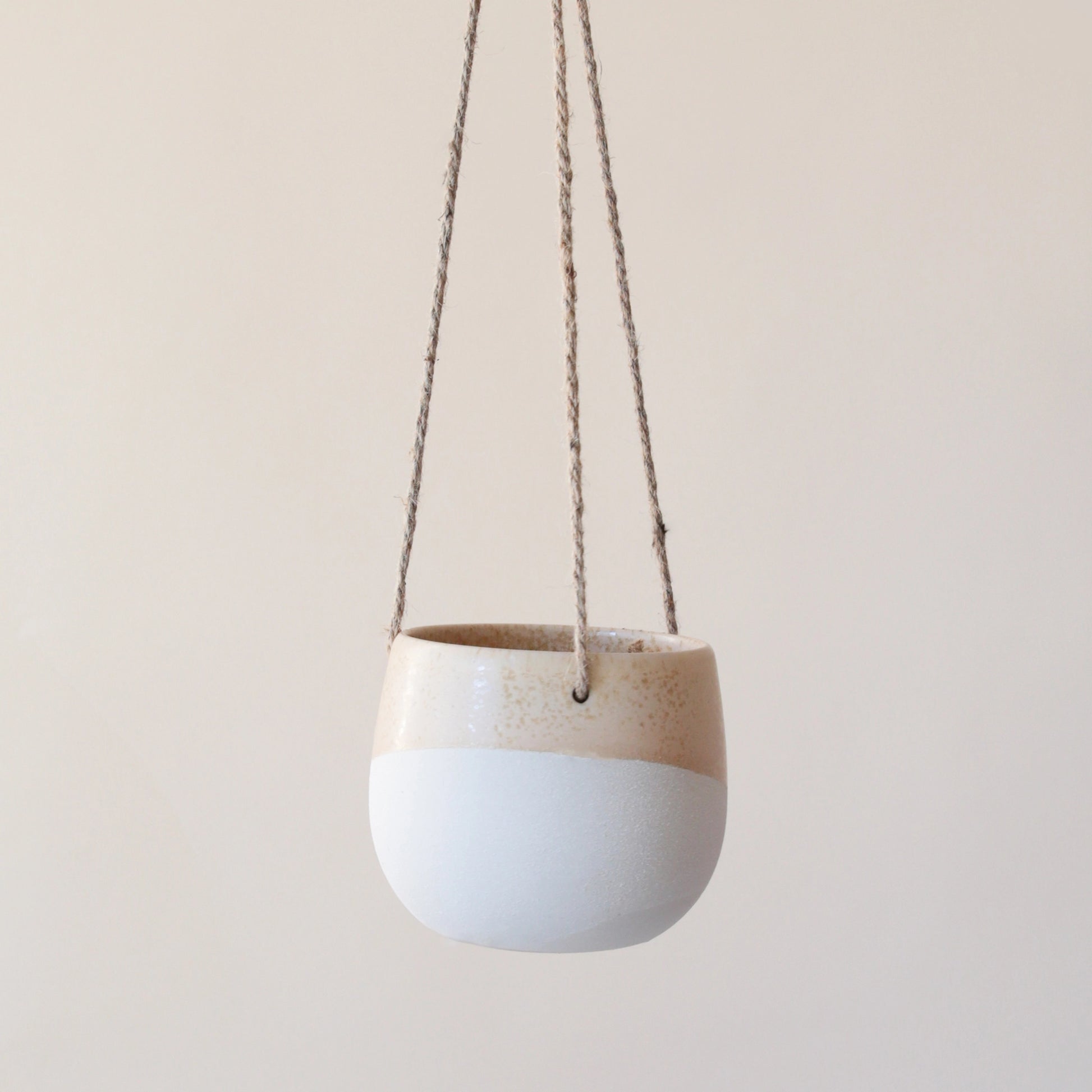 The smaller  neutral ceramic hanging planter with rounded sides and a flat bottom along with a half cream half tan design and three jute strings as the hangers.