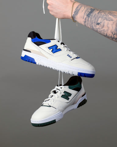 New Balance 550 trainers hanging from their laces. One in sea-salt white and angora green, the other in sea-salt white and blue.