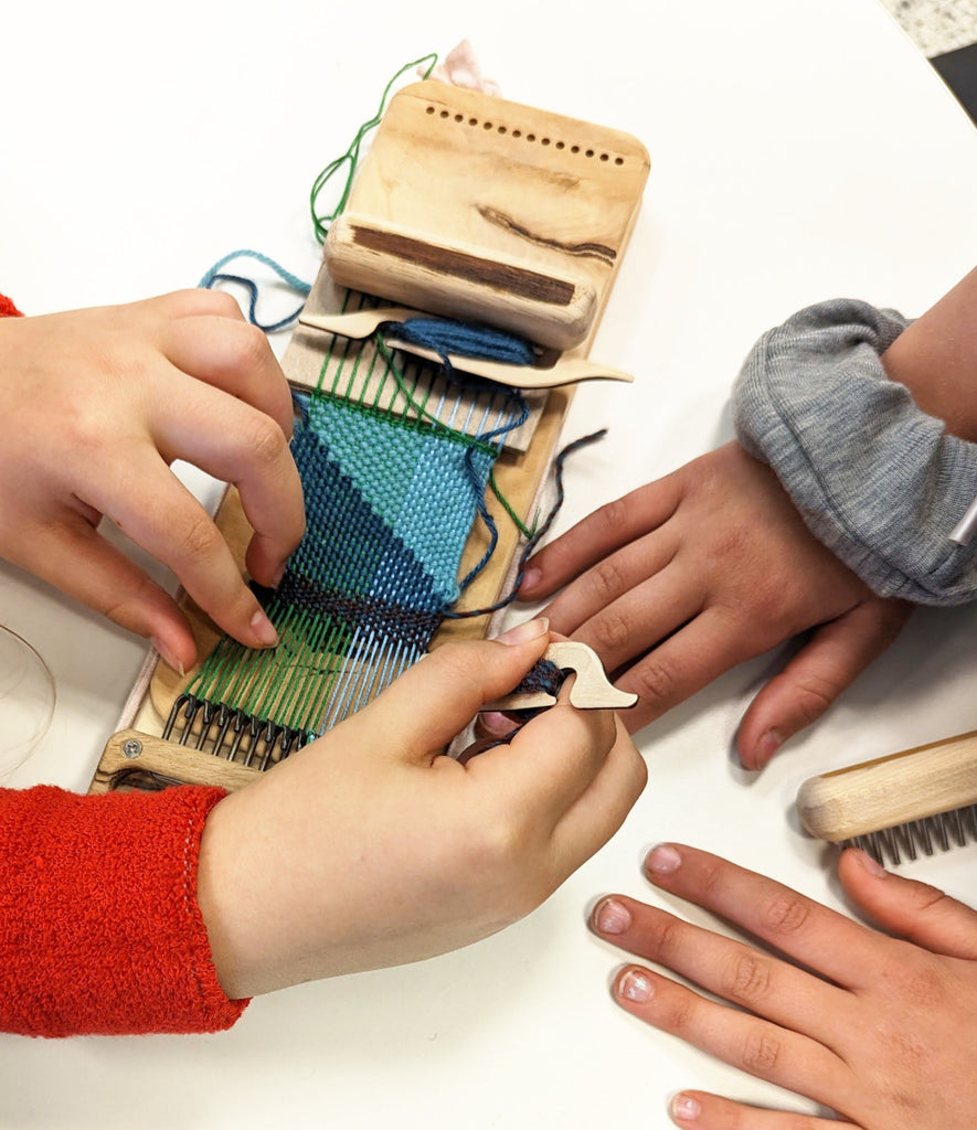 Two sets of pale hands work together to weave a miniature blue and green tapestry with the Swift Darning Loom.