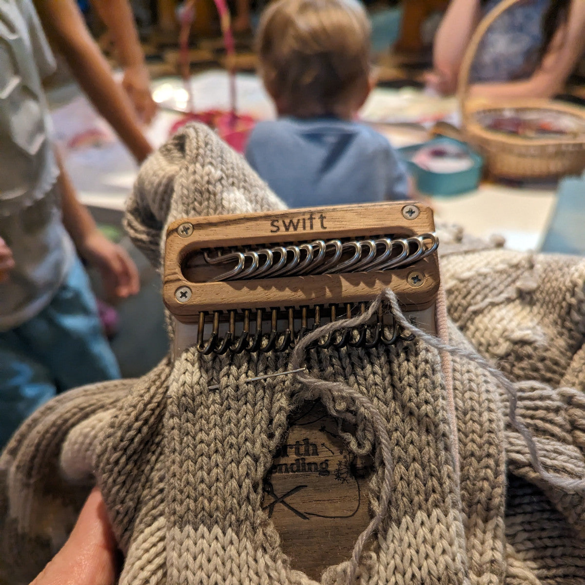 Swift Darning Loom showing off a holey sweater that's ready to mend, with lots of activity happening out of focus in the background as this mending project was taken on the go to church and errands.