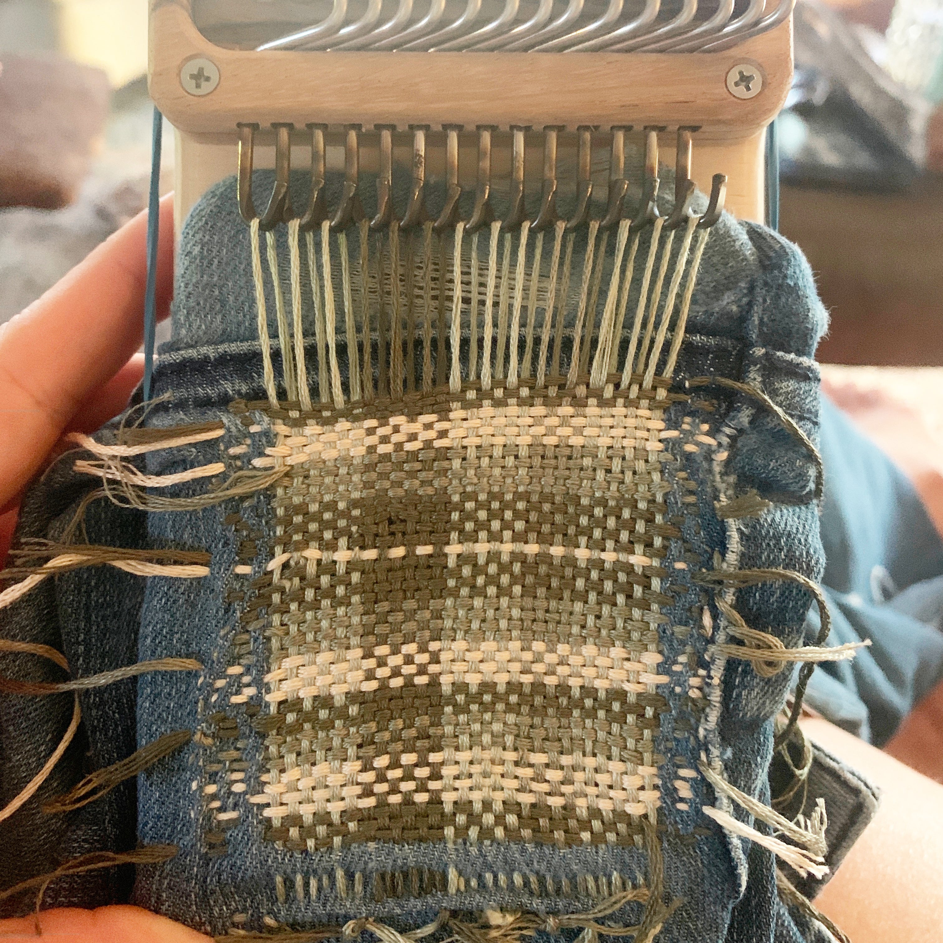 Swift Darning Loom in the process of weaving a large, multicoloured plaid patch on a pair of worn jeans.