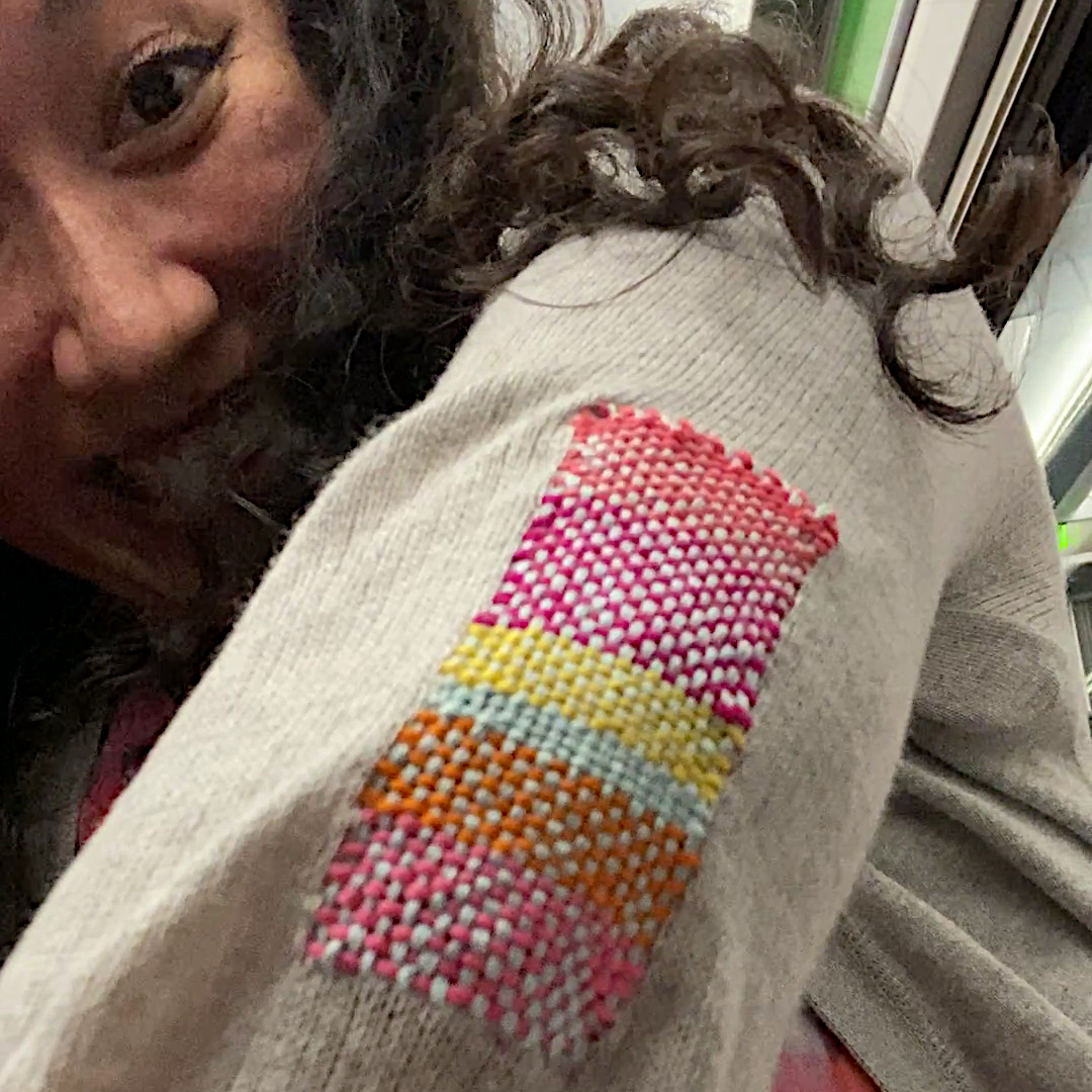 Athena of Meticulous Ink showing off a brightly coloured visible mending patch on a well-loved grey cardigan