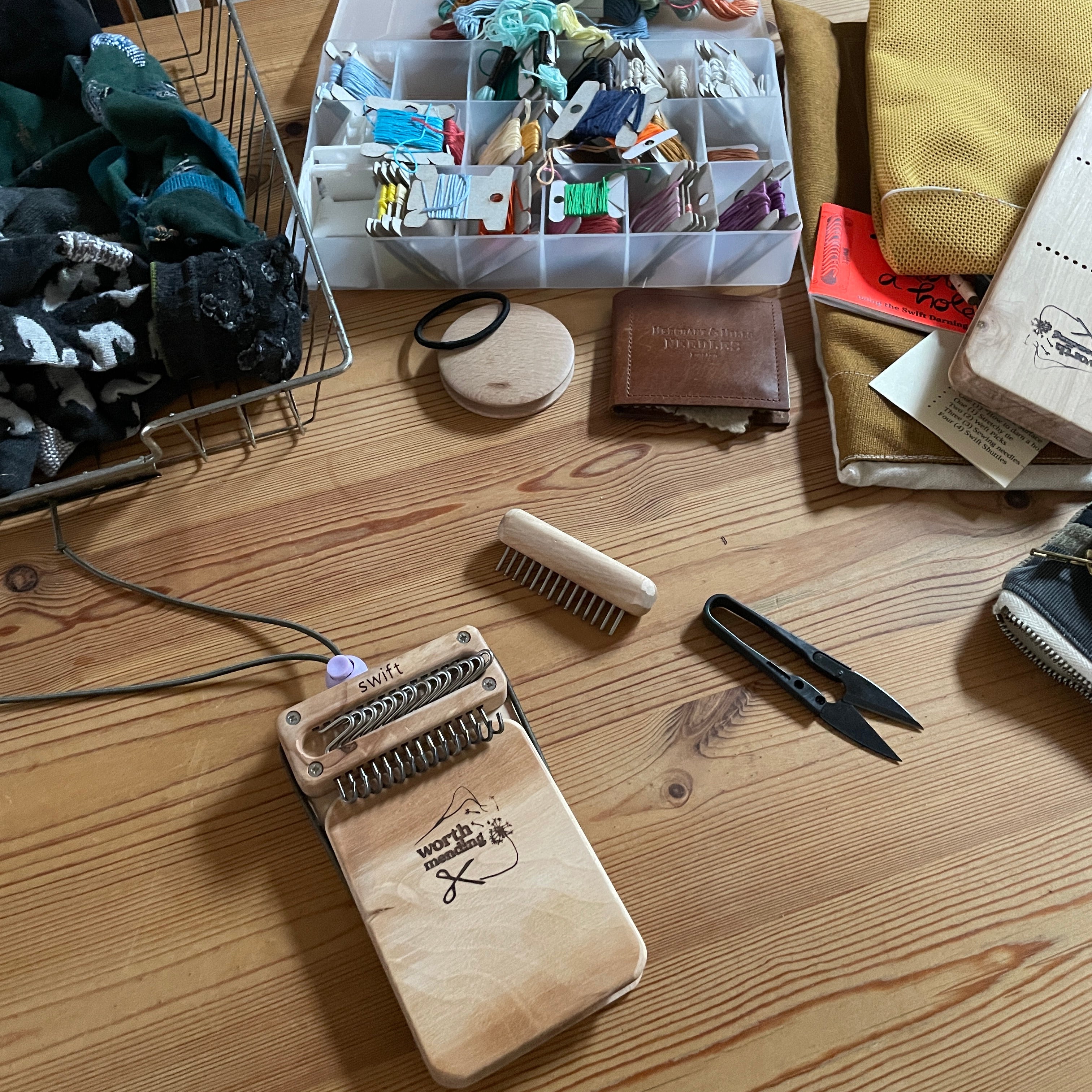 a wire basket full of mending, a plastic case of embroidery bobbins, and the contents of the Swift darning loom mending migration bundle are scattered over a wooden table.