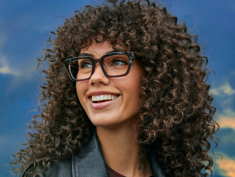 Woman with curly hair and glasses smiling against a blue sky background.