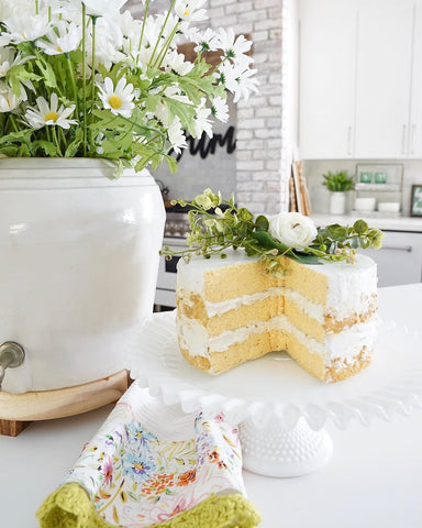 Faux cake with slice out on white cake stand with large vase of daisies