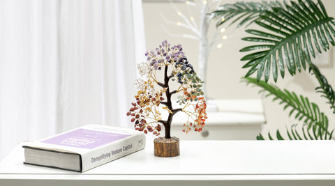 "A colorful gemstone tree decor with amethyst, quartz, and other crystals intricately arranged to mimic foliage sits next to a large book titled 'Demystifying Venture Capital' on a white table, with a potted tropical palm plant in the background, creating a serene office decor vibe."