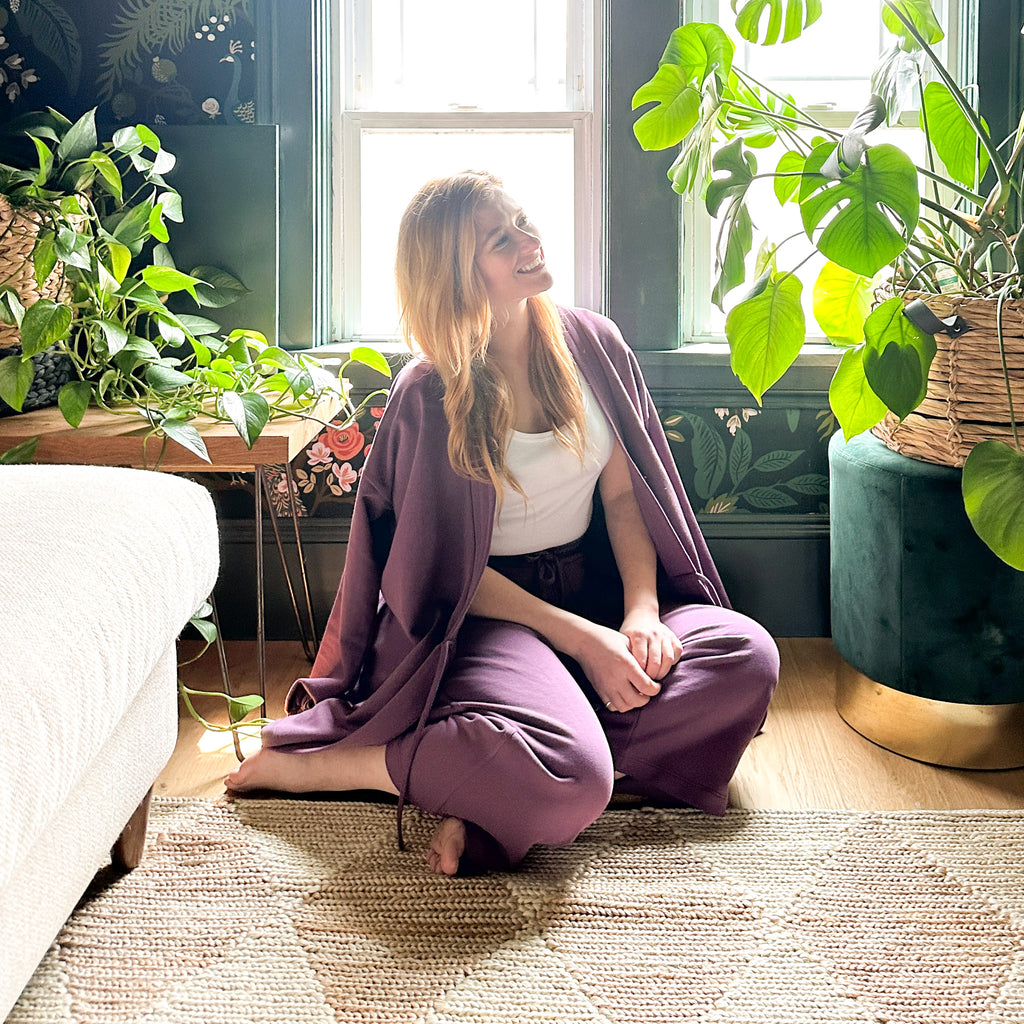 Photo of Rebekah Higgs aka DIY Mom sitting on the floor at her home with a bright sunny window behind her and surrounded by plants 