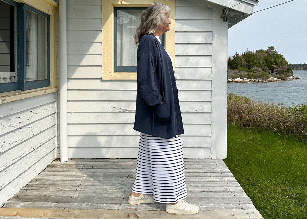A photo of a women wearing the Tokyo Coat in Deep pea and the Sailor Pant in Deep Sea Stripe standing in front of a white house with the ocean in the background 
