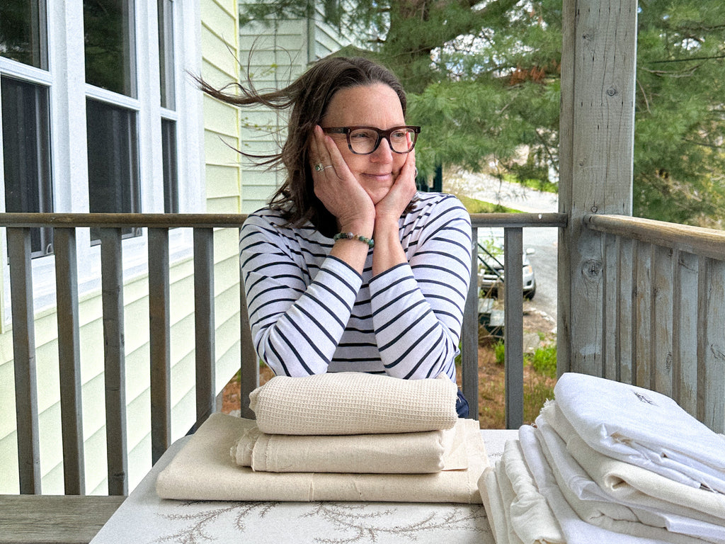 A photo of Lauren Chenoweth sitting on her deck with the breeze in her head with her organic fabrics organized in piles in front of her 