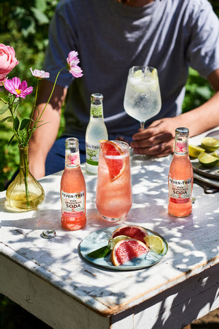 A nice garden table with Pink Grapefruit Sodas bottles served on iced glass and a person on the background holding a fresh glass