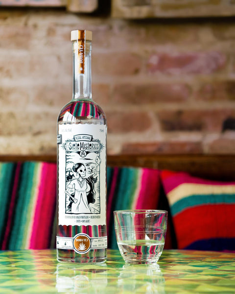 a bottle of Siete Misterios Doba-yej Mezcal and a half filled glass on a table with colored pillows in background in a room with visible old red bricks