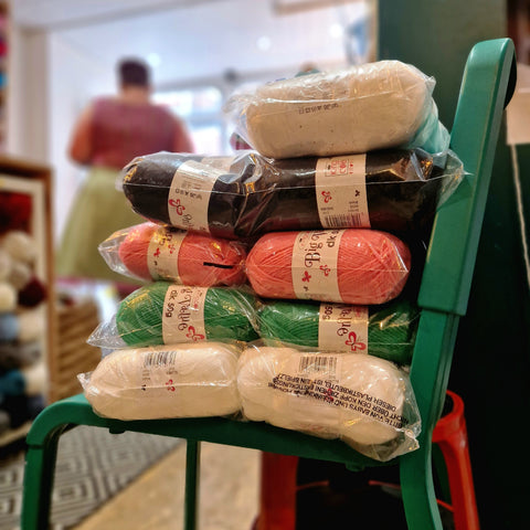 A stack of different colours of yarn on a green chair, with the shop in the background. From the bottom up, the yarn packs are white, green, pink, black and then white again.