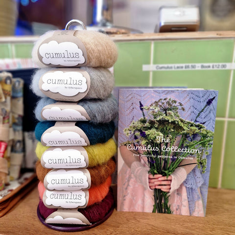 On the left is a stack of fuzzy yarn in all different colours. On the right is a pattern book with flowers on the front that says "The Cumulus Collection". The background is green tiles.