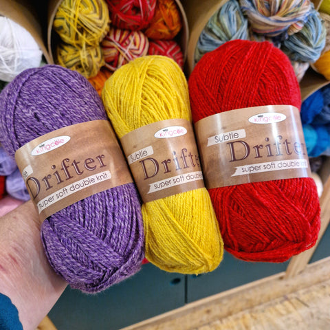 An arm holds three yarn balls in purple, yellow and bright red. The ball band says "King Cole Subtle Drifter Double Knitting" and is light brown. Shelves of yarn appear in the background.