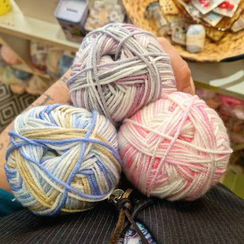 Three balls of yarn in multicoloured pastel shades are held against a body, looking down on the triangle of yarn.