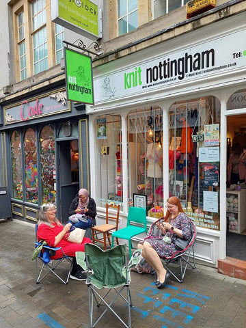 A group of three women sat on camping chairs knitting and crocheting outside of the wool shop - it looks damp and drizzly. 