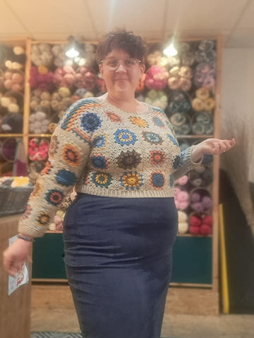 Eleanor, a fat, white woman wears a crochet jumper with deep jewel tone granny squares and a beige background.