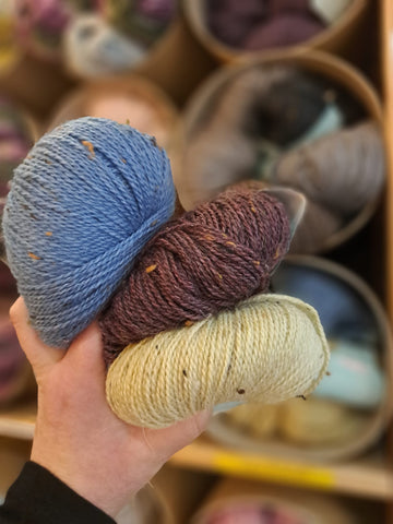 Three doughnut balls of yarn held in a white hand against a backdrop of brownish shelves of yarn. The balls in the hand are a bright denim blue, a deep aubergine, and a water-y wheat sort of yellow. All have beautiful flecks in. 