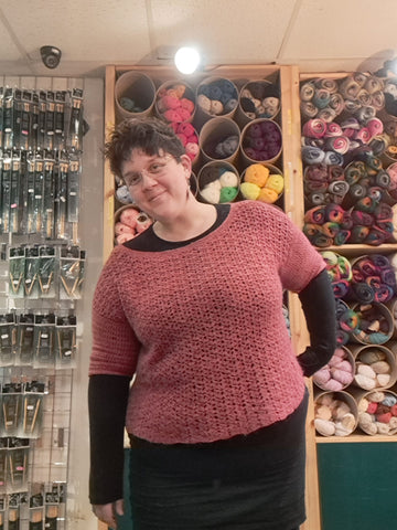 Eleanor, a fat white woman with short brown hair wears a red, crocheted jumper over a black outfit. She's standing at an awkward angle but smiling and she's stood in front of shelves of yarn. 
