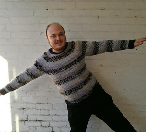 Dr Chris, a white man with very little hair and a beard stands at a funny angle with his arms outstretched in front of a painted white, brick wall. He's wearing a fairisle jumper in shades of blue. 