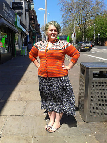 Eleanor, a fat white woman with grungy, greeny, bluey hair stands on a street with her hands on her hips. She's wearing a longlish, black summer dress with flip flops and a bright orange cardigan with a black and white fairisle yoke. 