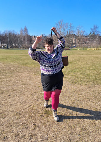 Eleanor, a fat, white woman with short brown hair walks towards the camera with her arms up in the air putting her cross body bag on. She's wearing a beige, navy, blue and purple fairisle jumper, a black skirt and bright pink tights. There's a lot of energy to the photo. 