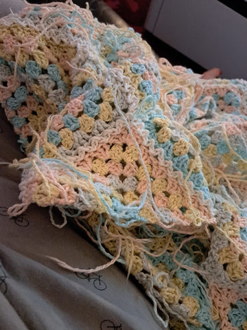 A pile of crochet on a lap. It's granny squares in pastels but not obvious what it is. There are lots of ends. 