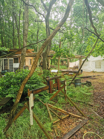 A soggy looking forest with a large wooden structure in the foreground. It's delicate looking, there's not a lot of wood but they're long pieces fashioned together with string. I think it makes some sort of turning effect so that wood can be shaped. There are ramshackle huts in the background. 