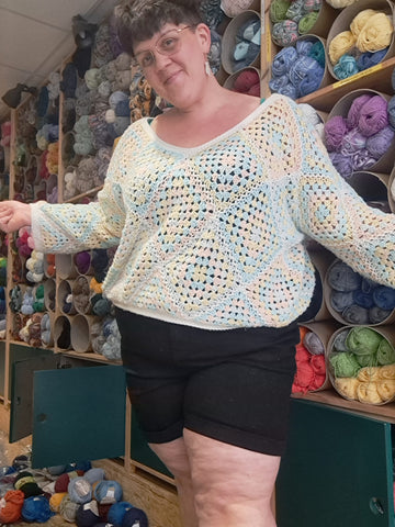 Eleanor, a fat white woman with short brown hair poses for the camera wearing black shorts and an oversized, crochet jumper in pastel shades. The shop behind her is a mess.