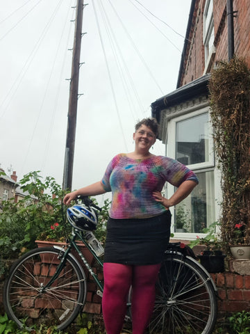 Eleanor, a fat, white, woman, with short, brown hair stands in front of a bike in front of a messy garden. She's wearing bright pink tights, a black skirt and a multi coloured crochet jumper. Hand on her hip and smiling.