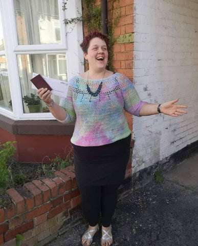 Eleanor, a fat, white, woman wears a multi coloured, crochet jumper in front of a brick house. She's got a black skirt on and a blue necklace and she has her arms spread wide.