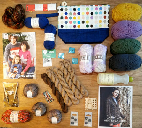 [ID: A flat lay of various yarns, pattern books, buttons, knitting needles and other knitting goodies on a brown table. It's very colourful. End ID.]
