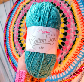 A disembodied white hand holds a blue ball of yarn against a crocheted mandala in vibrant colours. The ball band on the yarn says Cottontop DK