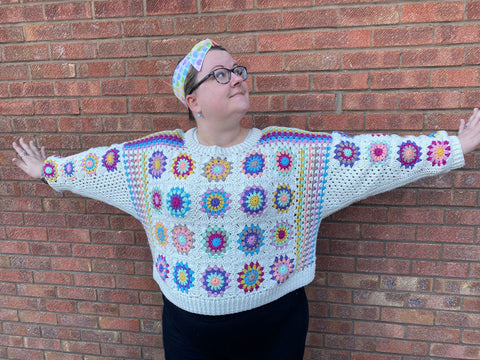 Rachel, a white woman with her hair in a pastel coloured scarf stands with her arms outstretched in front of a brick wall. She's wearing a granny square jumper in beautiful pastel shades with a creamy white edging 