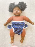A dark skined doll wearing a modern preflat cloth nappy with a dinosaur cover