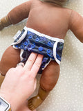 A dark skined doll wearing a modern preflat cloth nappy with a dinosaur cover