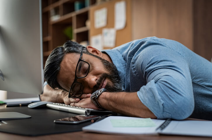 Mann schläft mit dem Kopf auf dem Schreibtisch und der Tastatur. Starke Müdigkeit aufgrund von zu wenig Bewegung.