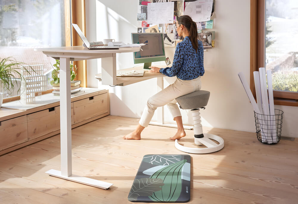 Frau sitzt an einem höhenverstellbaren Schreibtisch auf dem Aeris Swopper. Mehr Bewegung im Home-Office fördert die Gesundheit.