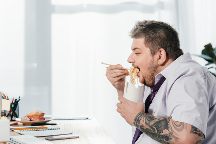 Übergewichtiger Mann isst sein Mittagessen am Schreibtisch. Bewegungsmangel und die Folge Übergewicht.