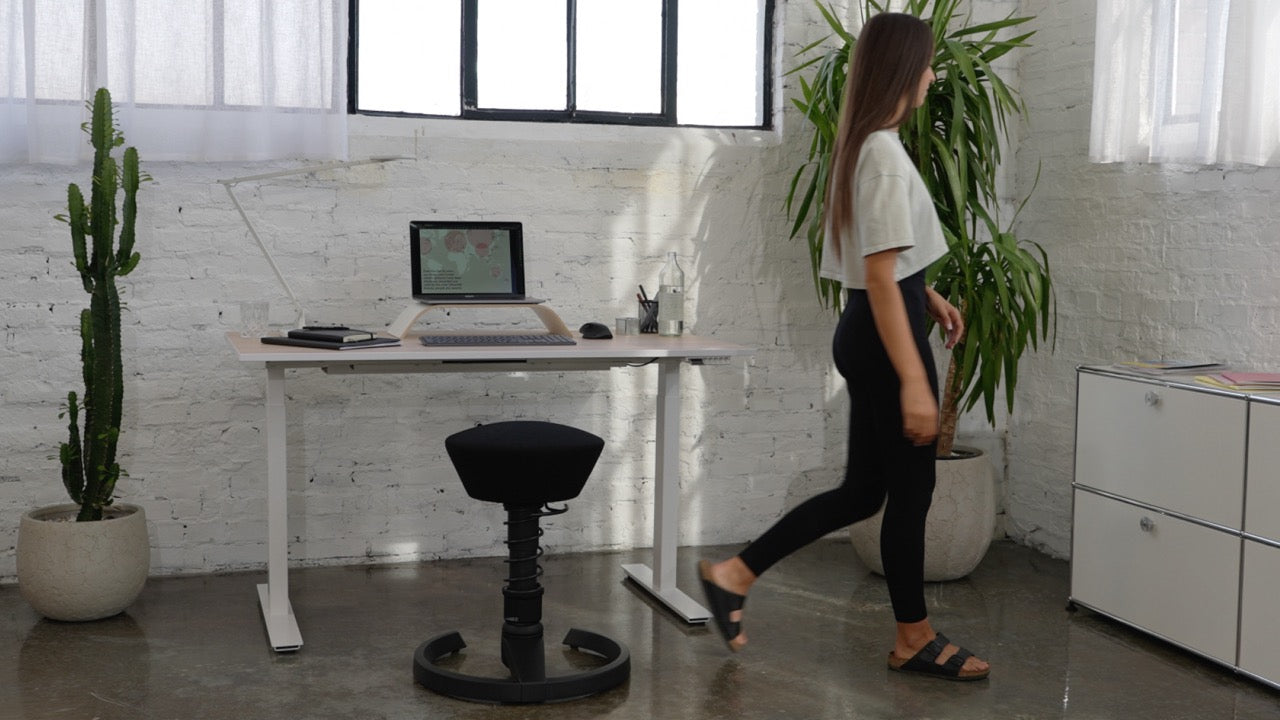 Vrouw op kantoor met het in hoogte verstelbare bureau Active Desk en bureaustoel Aeris Swopper, die voldoet aan de DIN-norm voor bureaustoelen met rugleuning en wielen.