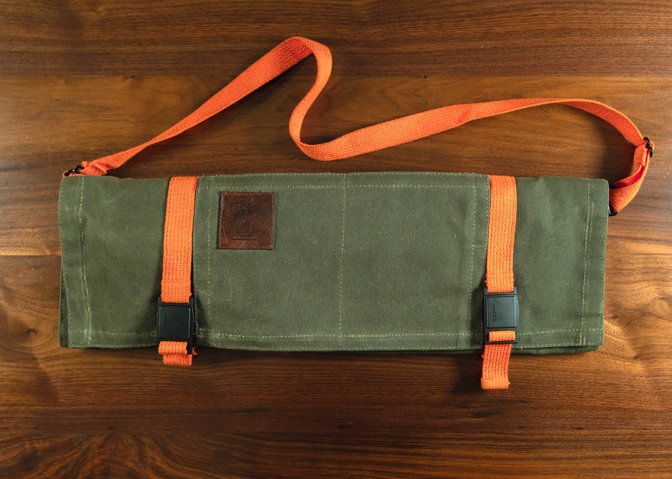 Cotton olive green knife roll protected by tex-wax with orange strapping displayed on a wooden surface. The Main Squeeze knife roll was manufactured in Minnesota by Craftmade Aprons.